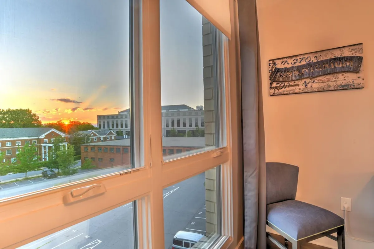 A comfortable chair sits near a window showcasing a vibrant sunset, with tasteful wall art creating a homely ambiance. The view hints at a scenic mountain backdrop, suggesting a connection to the natural beauty surrounding Asheville. The calm, reflective mood invites relaxation and family bonding at the end of an exciting day.