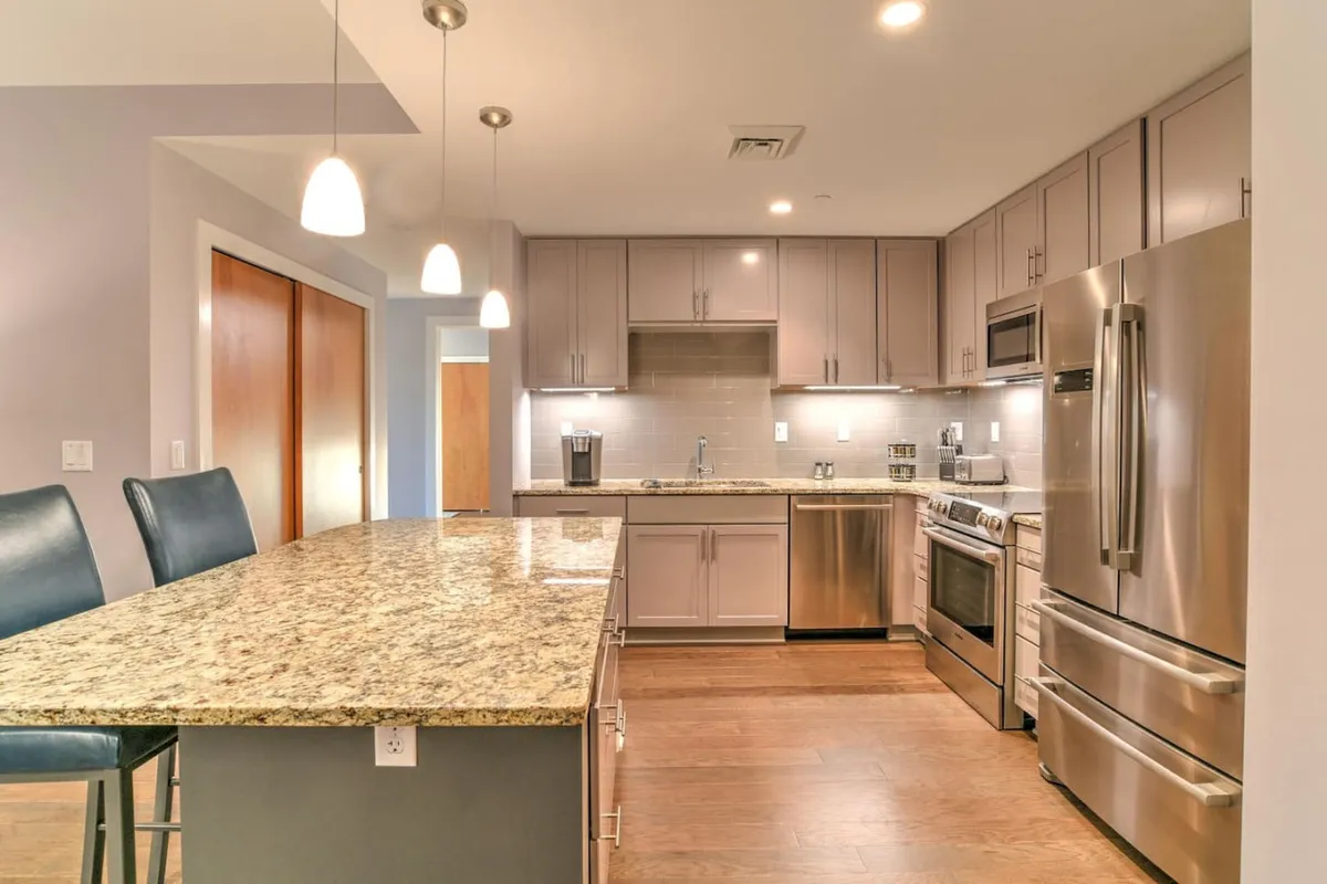 This image features a welcoming kitchen with a granite countertop, stainless steel appliances, and modern pendant lighting overhead. The scene suggests a space perfect for cooking, entertaining, and gathering, with ample prep areas and high-end finishes.