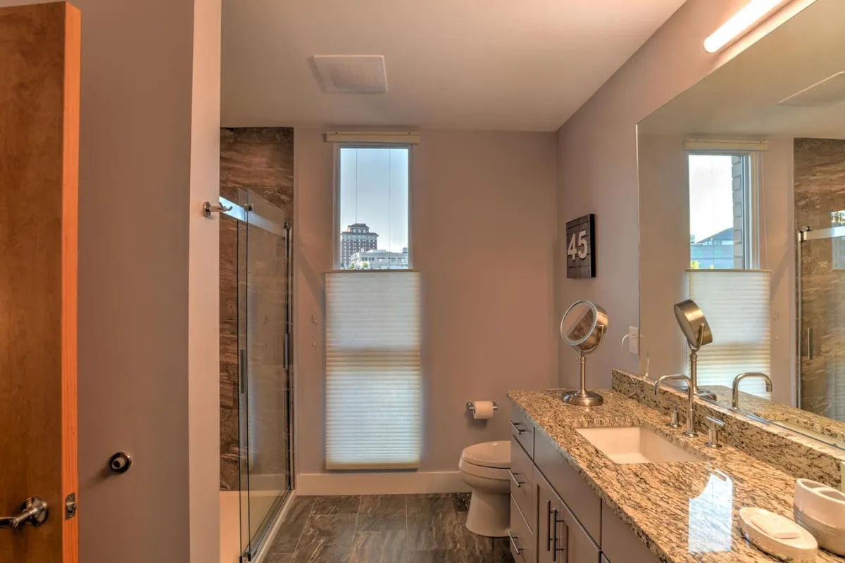 A modern bathroom with a granite countertop, a large mirror, a glass shower enclosure, and a window that offers a city view. The space feels inviting and serene, perfect for self-care routines and relaxation.