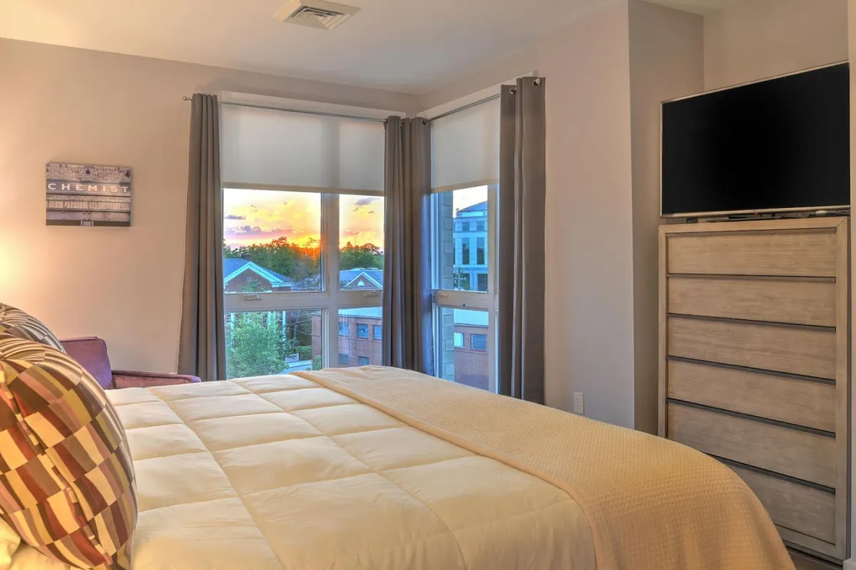 A cozy bedroom featuring a bed with patterned pillows, a window offering a serene sunset view, a dresser, and a television. The scene exudes warmth and coziness, inviting quiet relaxation and comfort.
