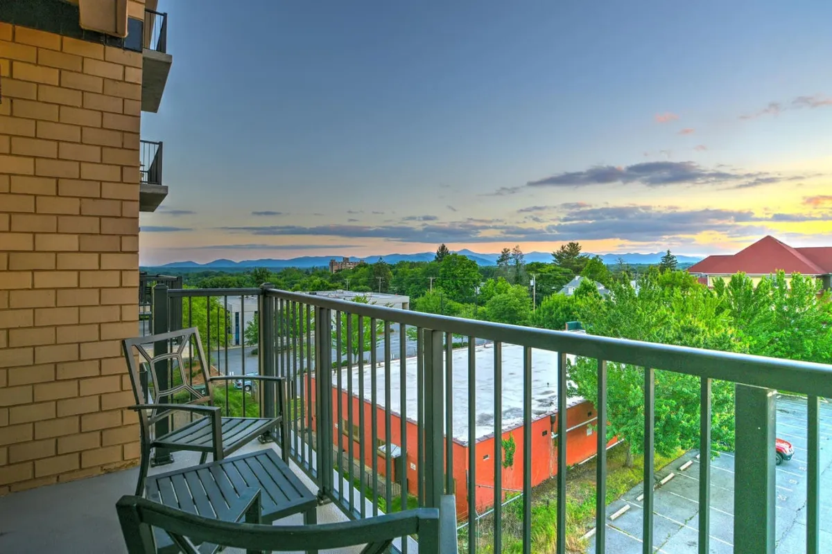 A wider view from the balcony, featuring a sturdy railing, stylish outdoor seating, and a panoramic vista of green trees, mountain ranges, and the vibrant sunset sky above Asheville’s urban landscape. The scene exudes serenity and possibility for gatherings or solo moments.