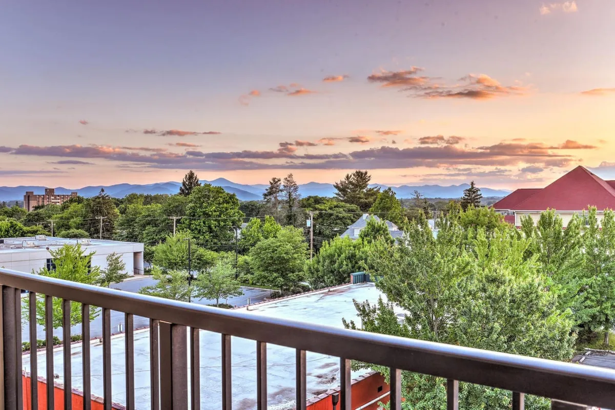 An elevated cityscape with green tree canopies, urban rooftops, and the Blue Ridge Mountains under a sunset sky. The perspective suggests the proximity to both city amenities and nature, as well as the potential for vibrant evenings spent with friends appreciating the view.