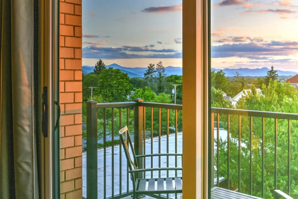 The photo features a balcony with a railing looking out over green trees, distant mountains, and a glowing sunset sky. The setting suggests a peaceful moment, ideal for relaxing together with a scenic mountain view.