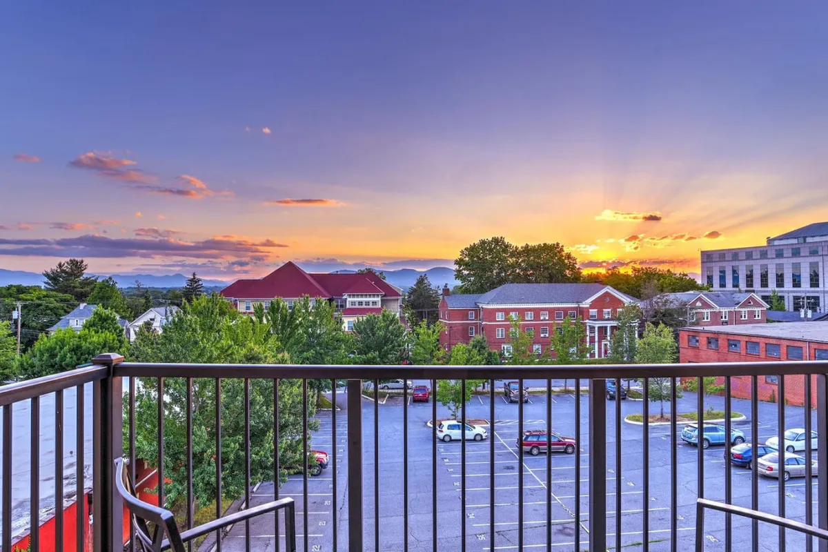 This image captures a tranquil balcony scene at sunset, with a clear view of the mountains beyond the railing. The sky glows with warm colors as the sun dips behind the peaks, suggesting a perfect spot for relaxing and socializing after a day of city adventures.