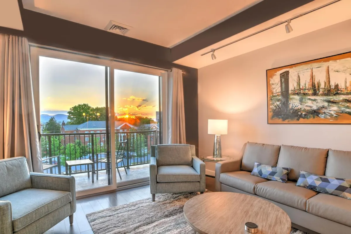 This image showcases a sun-drenched living area with expansive windows framing a breathtaking sunset over Asheville. Plush seating invites guests to relax, while tasteful artwork and a modern coffee table complete the cozy, upscale ambiance—perfect for unwinding or sharing memorable moments.